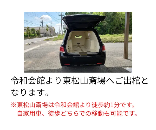 令和会館前の霊柩車の後方ハッチが空いてる写真。後方には東松山斎場の入口が写っている。
テキスト：令和会館より東松山斎場へご出棺となります。
※東松山斎場は令和会館より徒歩約1分です。自家用車、徒歩どちらでの移動も可能です。